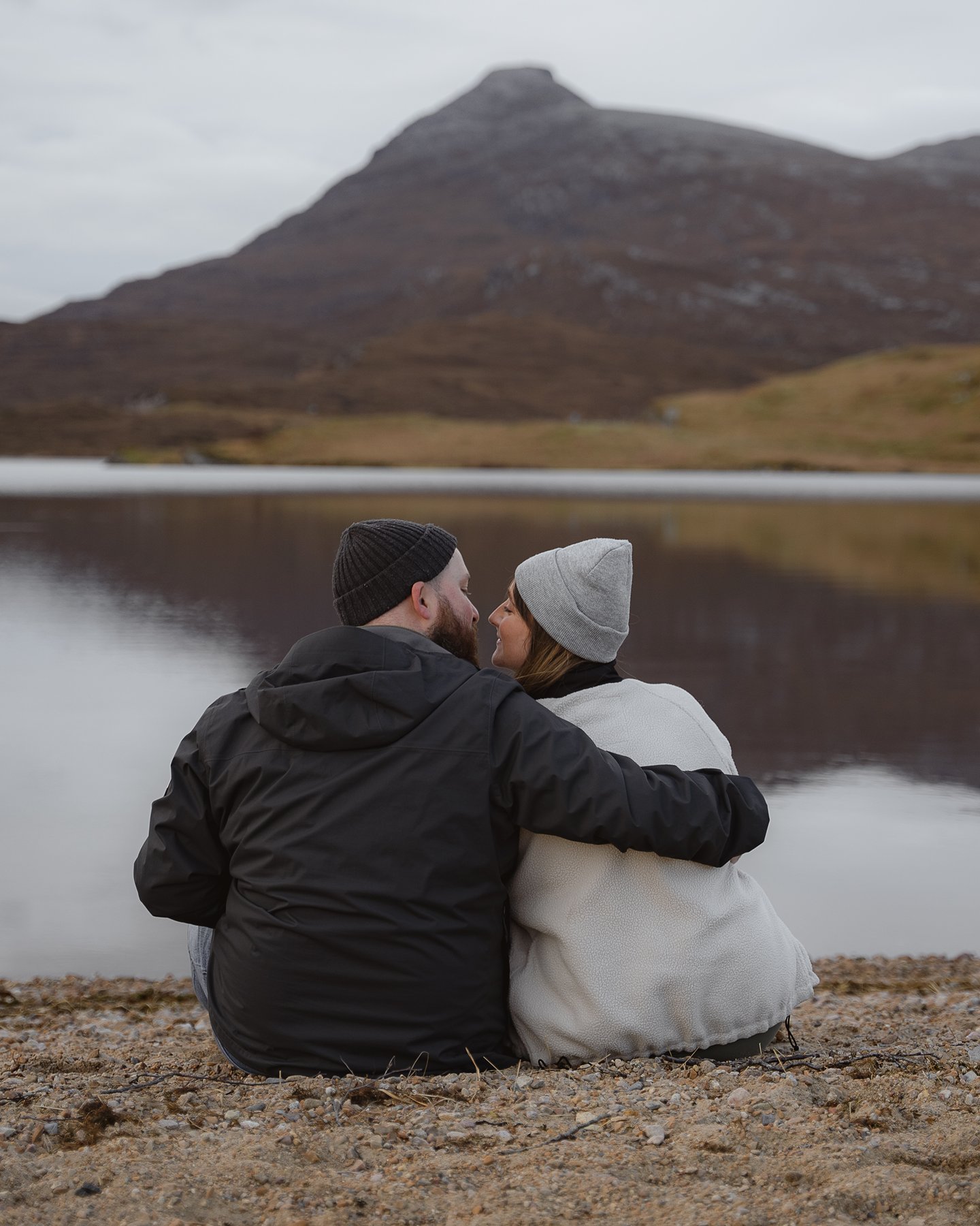 Couple kissing by Loch Assynt in Scotland with mountain backdrop, sitting on ground by the water.