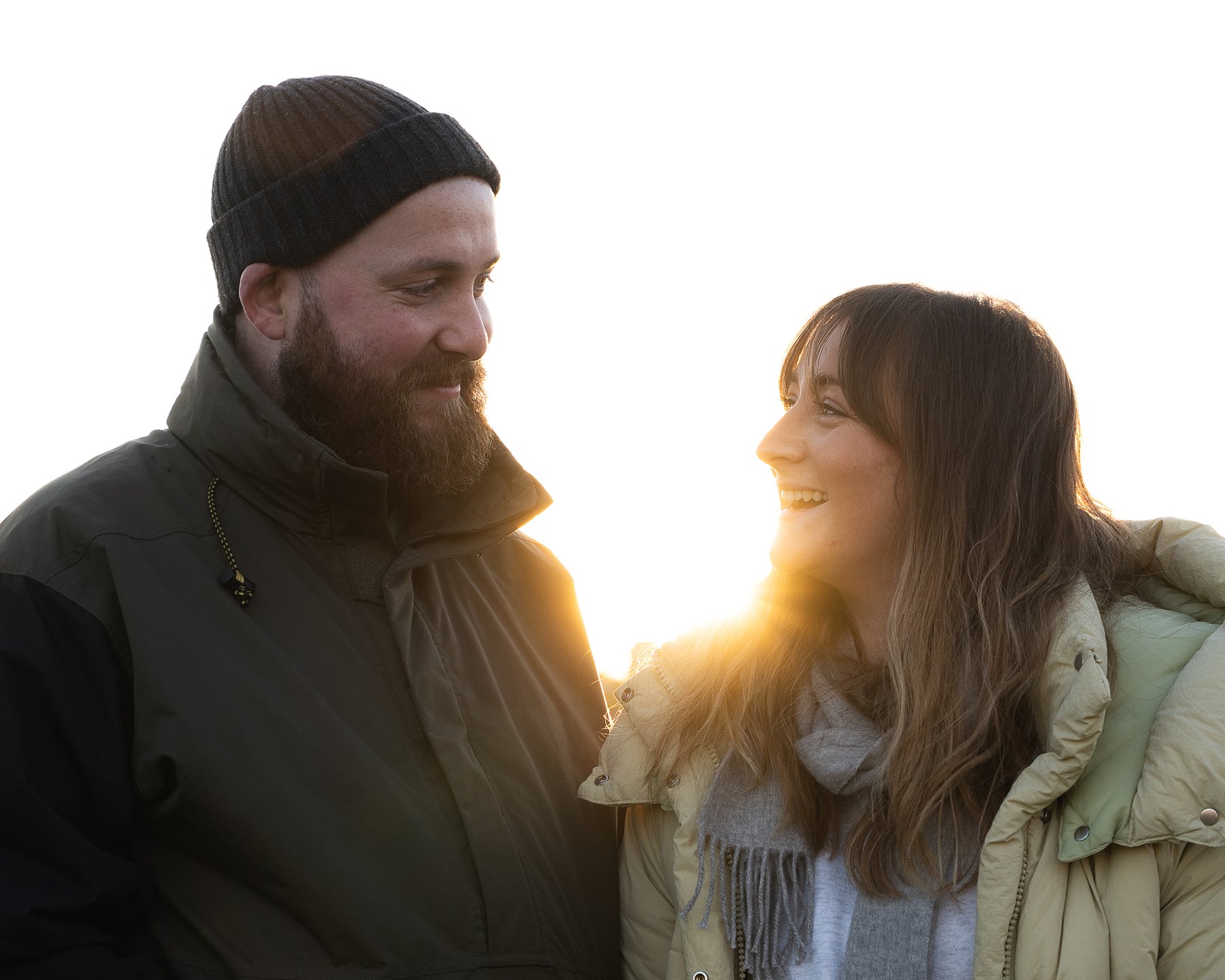 Couple in warm jackets smiling at each other outdoors, with sunlight shining between them.