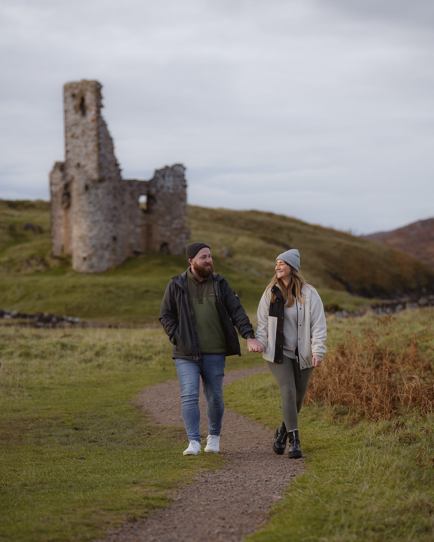 Smiling couple holding hands walking on path with historic stone castle ruins in Scottish Highlands.