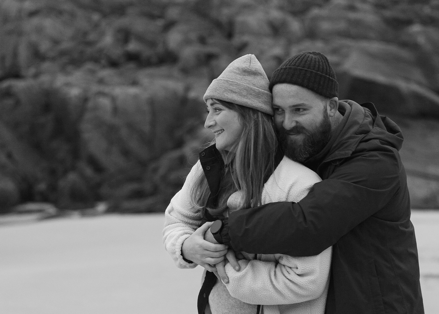 Couple cuddling on beach in winter clothing with knit hats, both smiling as they look off into the distance