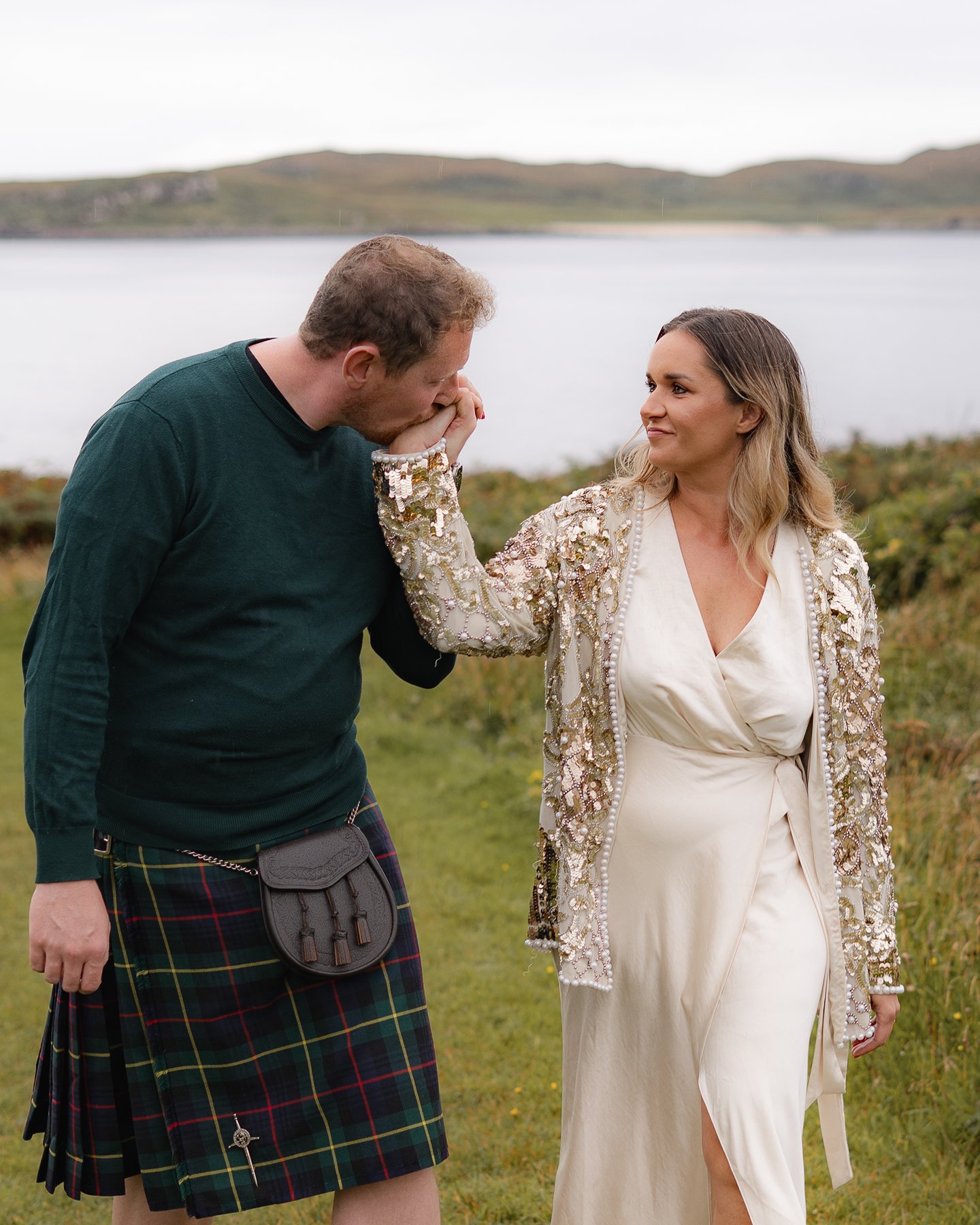 Couple in Scottish Highlands by the loch, groom in traditional kilt kissing bride's hand in white dress.