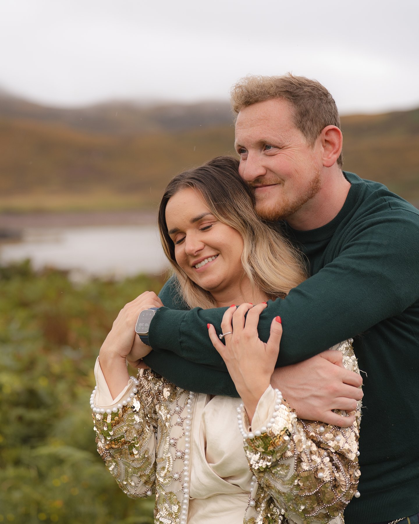 Couple embracing outdoors, with the man wrapping his arms around the woman as they smile in a scenic landscape.