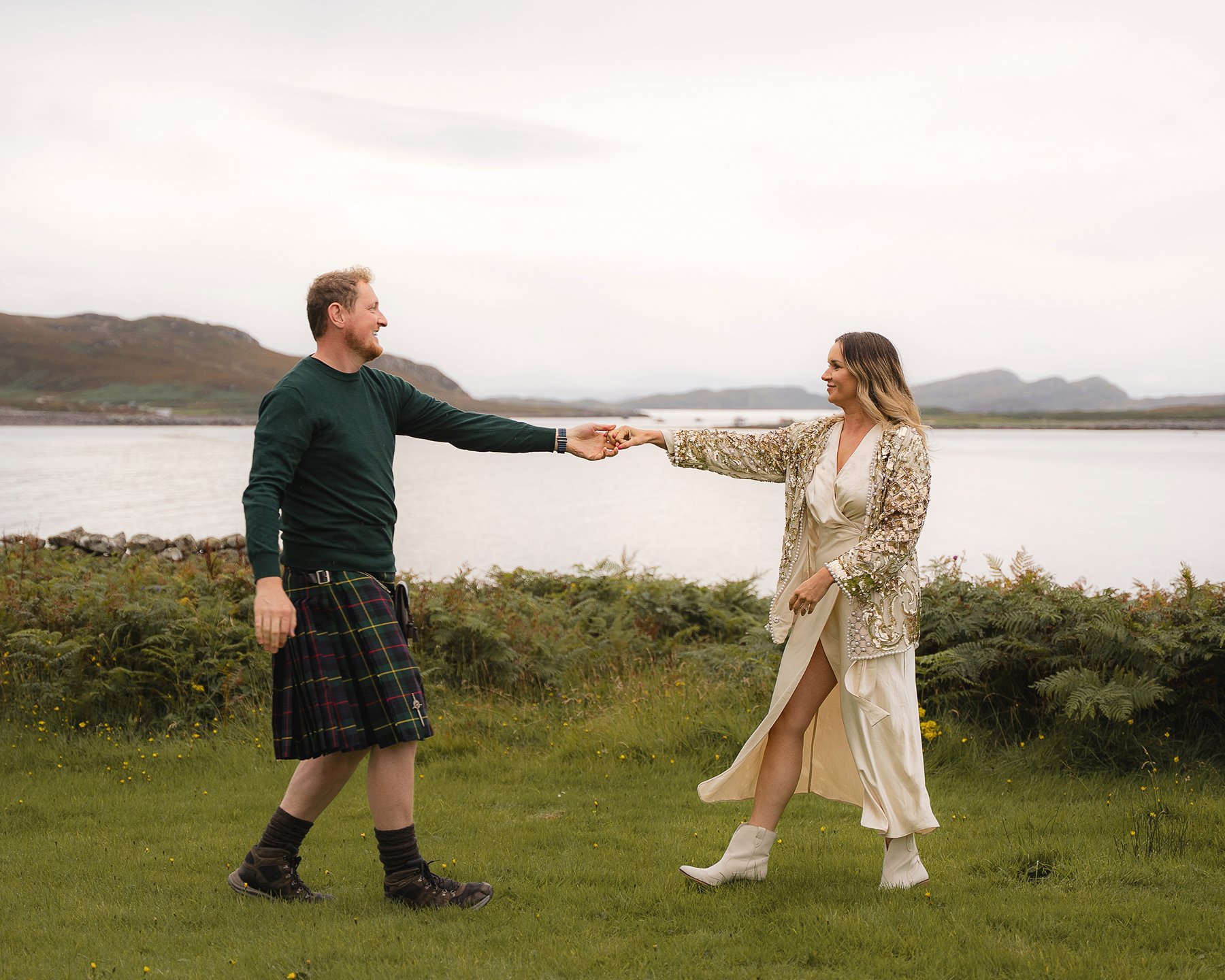 Couple holding hands and dancing outdoors by the water, man in kilt and woman in flowing dress with sequin jacket.