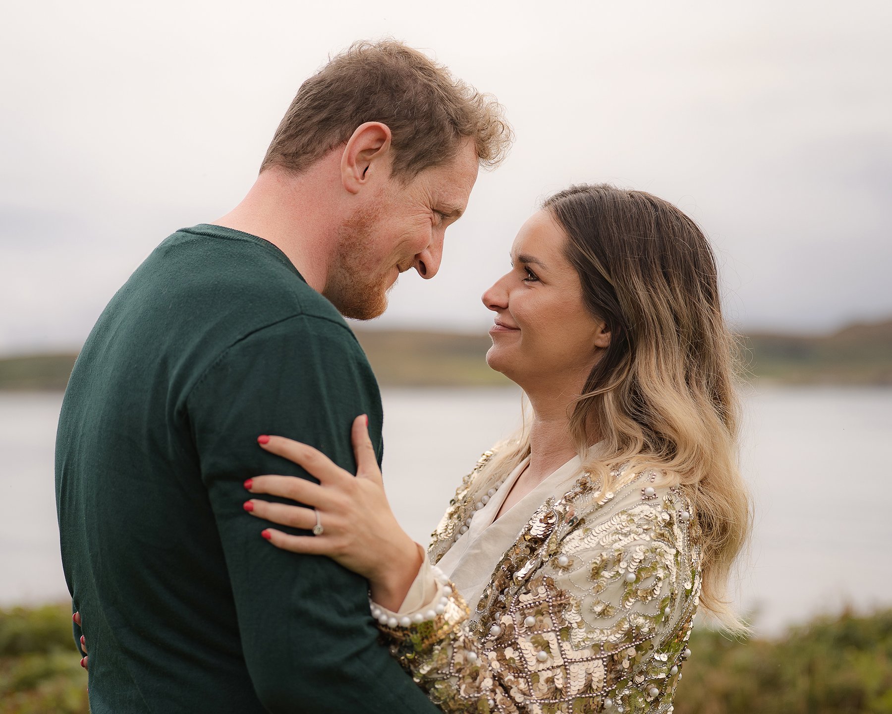 Happy engaged couple embracing by the loch in Scotland, woman wearing gold sequined top showing off her new engagement ring.