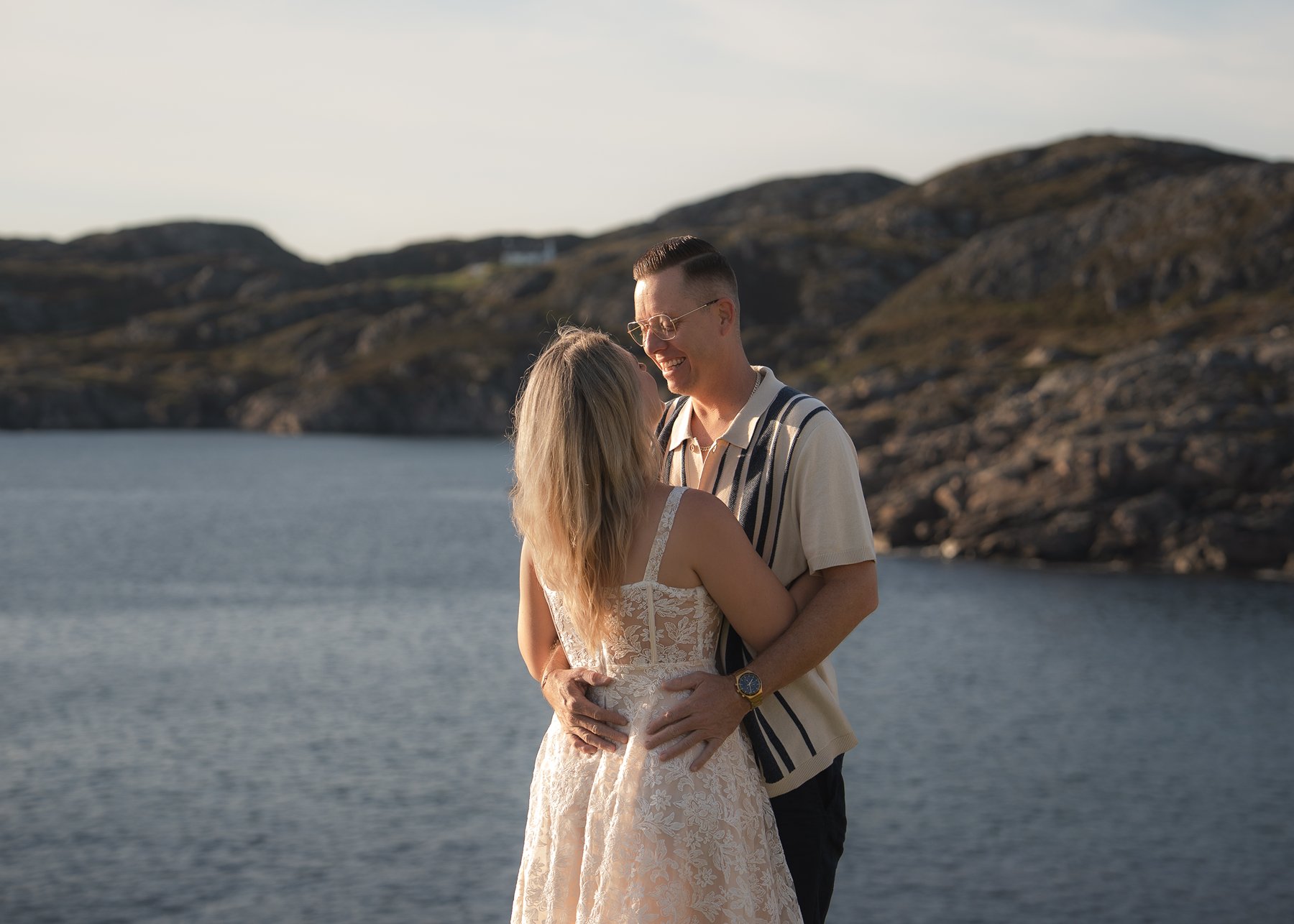 Smiling couple embracing by the sea, with rugged hills behind in the soft evening light.