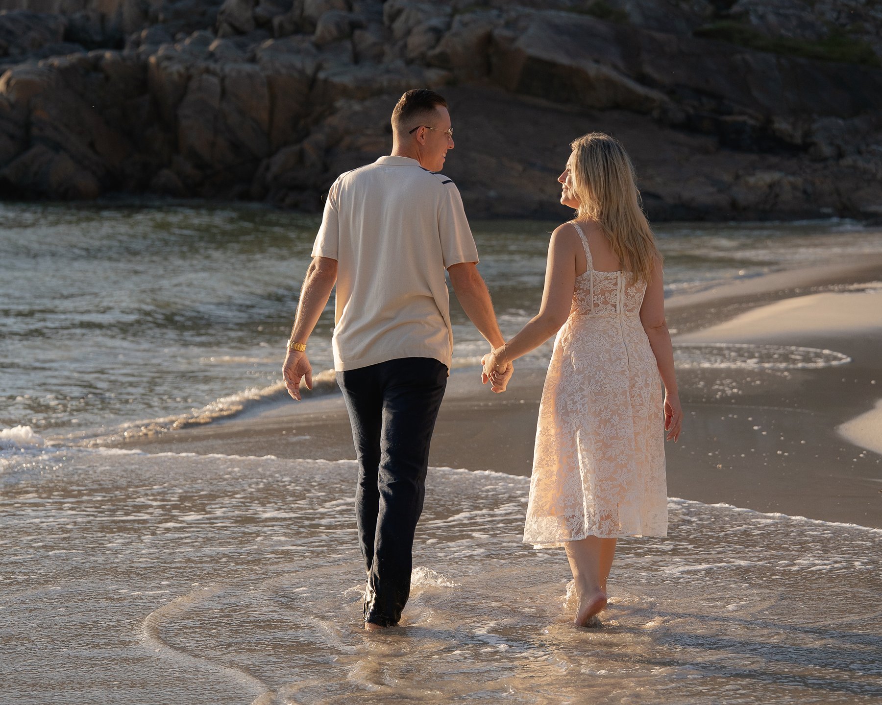 Couple holding hands and walking barefoot along the shoreline at sunset.