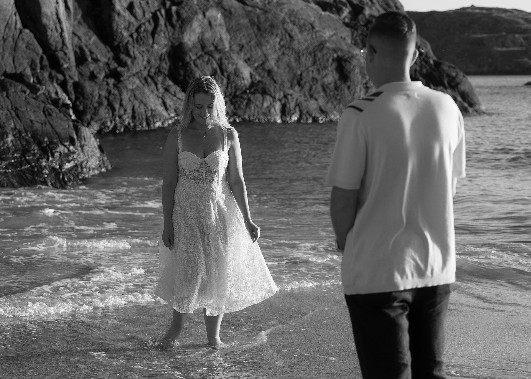 Man watching a woman in a lace dress as she walks barefoot in the shallow waves on a rocky beach.