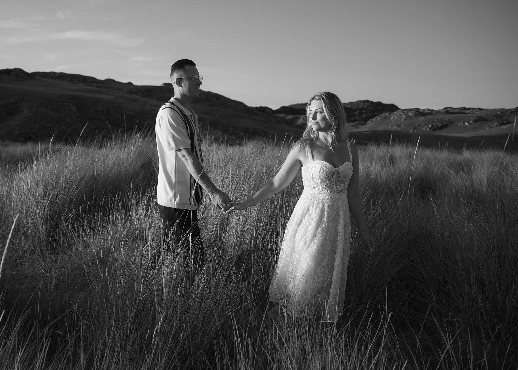 Engaged couple walking through tall Highland grass, woman looking back at him..