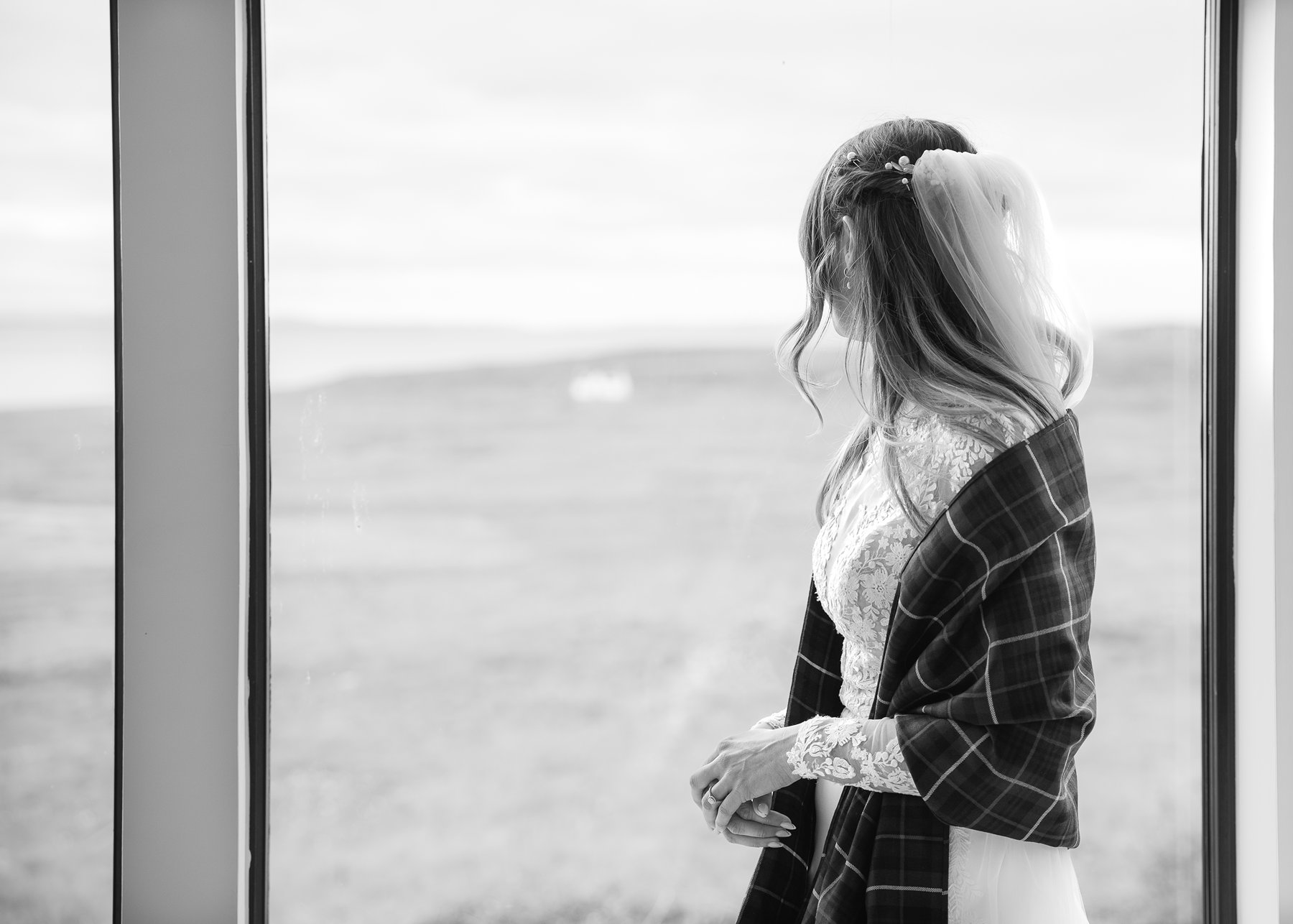 Bride in lace wedding dress with tartan shawl looking out window on wedding day.