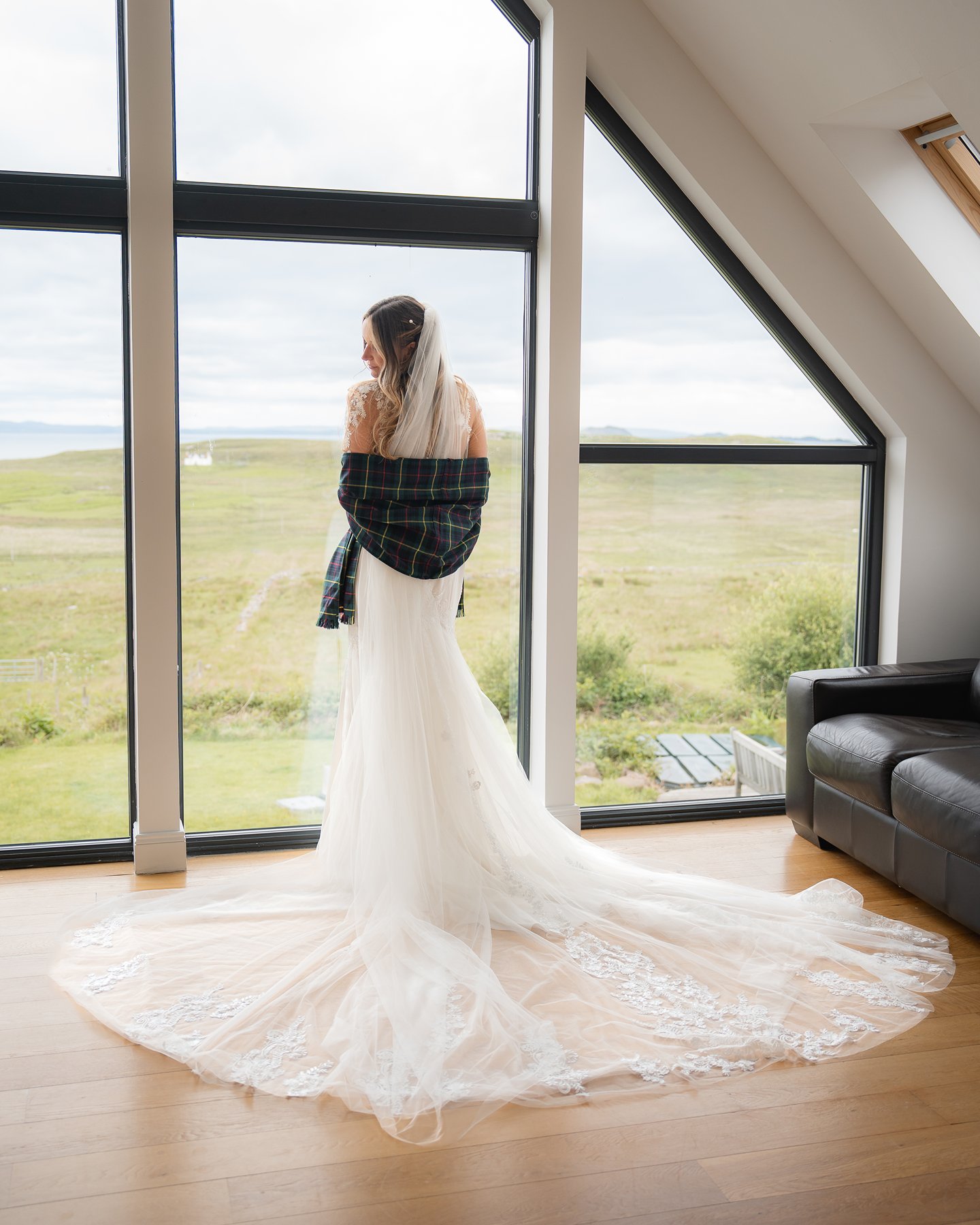 Bride in a white wedding dress wrapped in tartan shawl, standing by large window overlooking Scottish countryside.