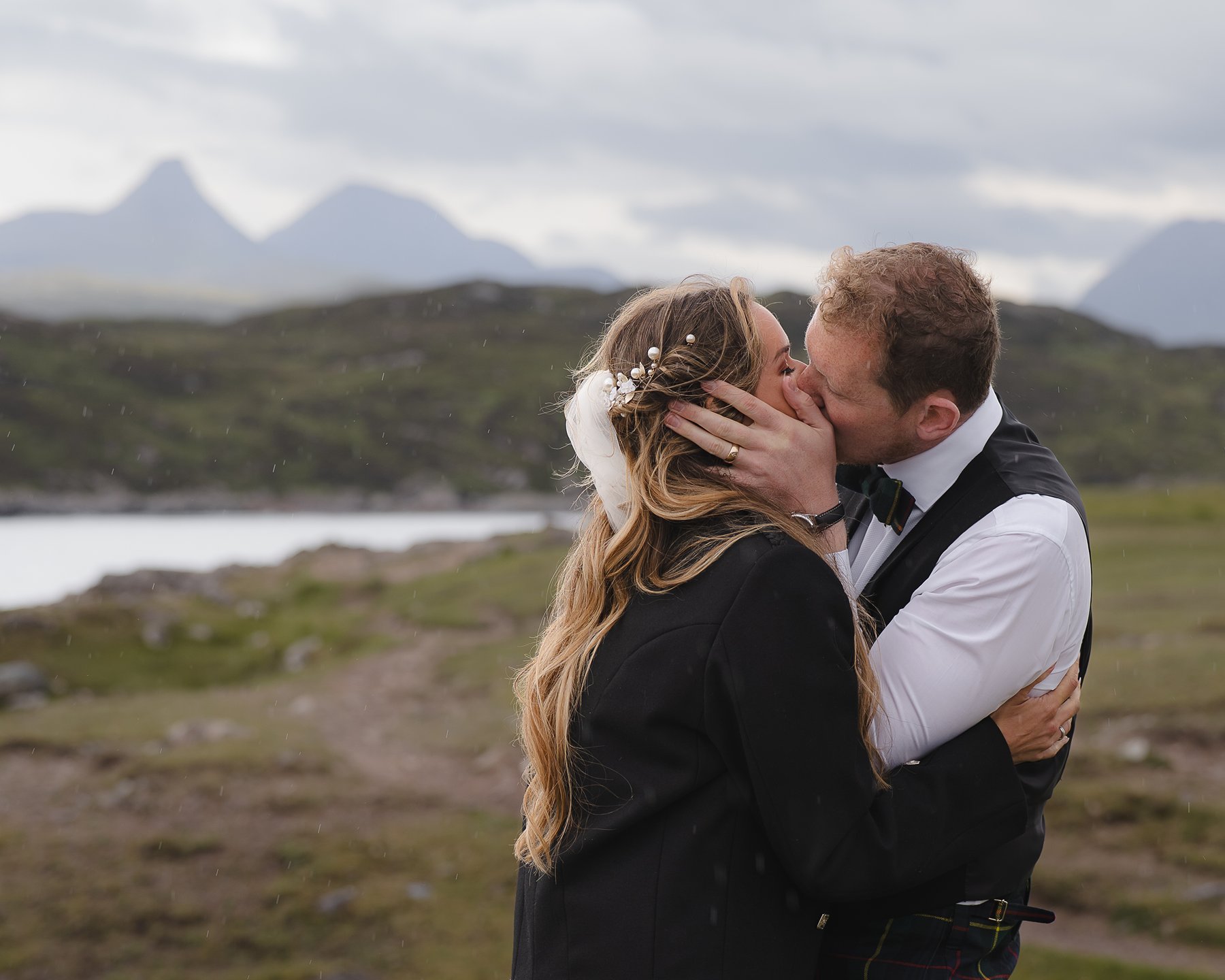 Married couple having a first kiss outdoors on their wedding day, with mountains and a sea in the background.
