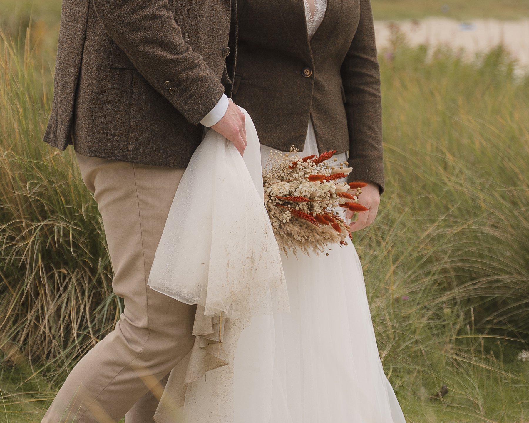 Close-up of Groom lifting bride’s dress while she holds a rustic dried flower bouquet.