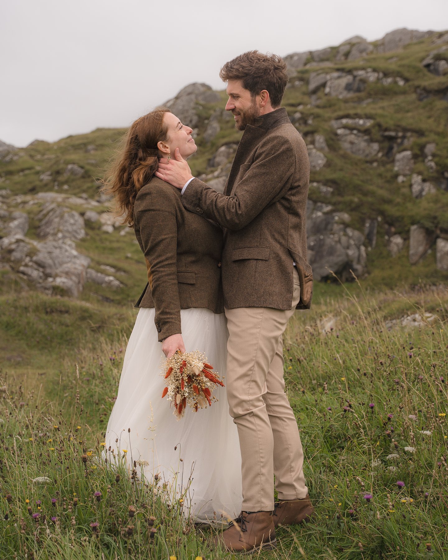 Smiling couple in tweed outfits standing in a grassy hillside, holding a bouquet.