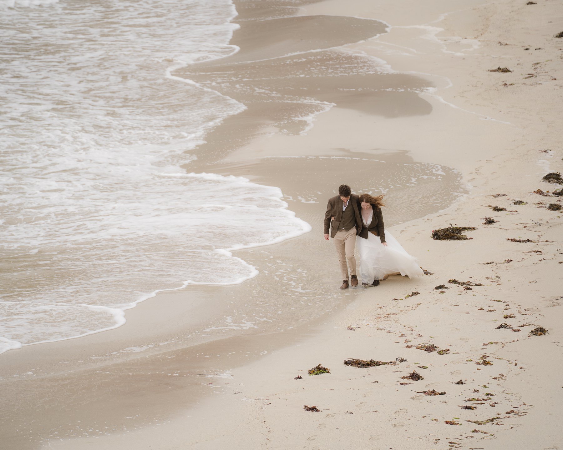 Bride and groom walking together on sandy beach with scattered seaweed and ocean waves washing ashore.