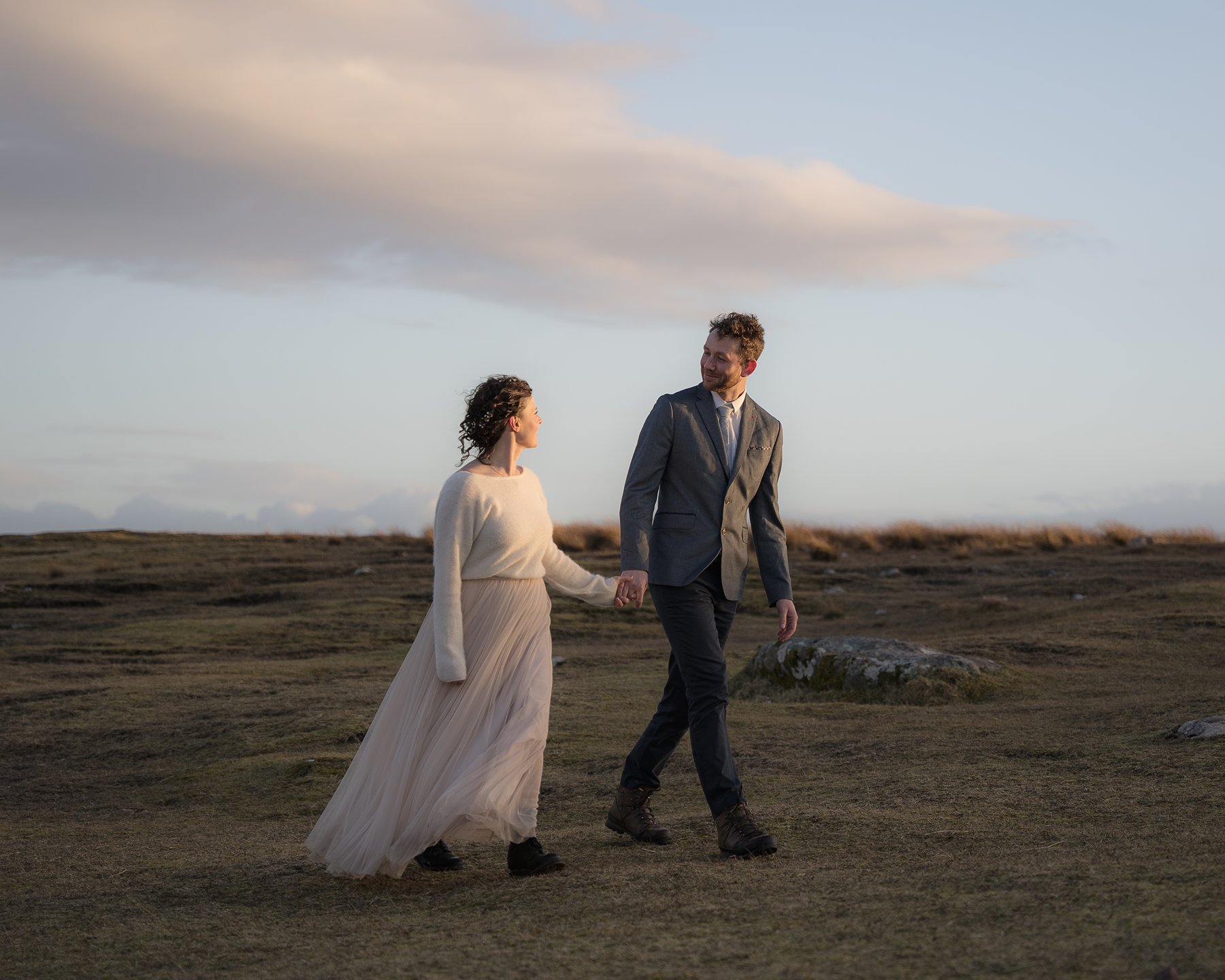 Newlyweds holding hands, walking across open moorland at sunset during their Scottish elopement.