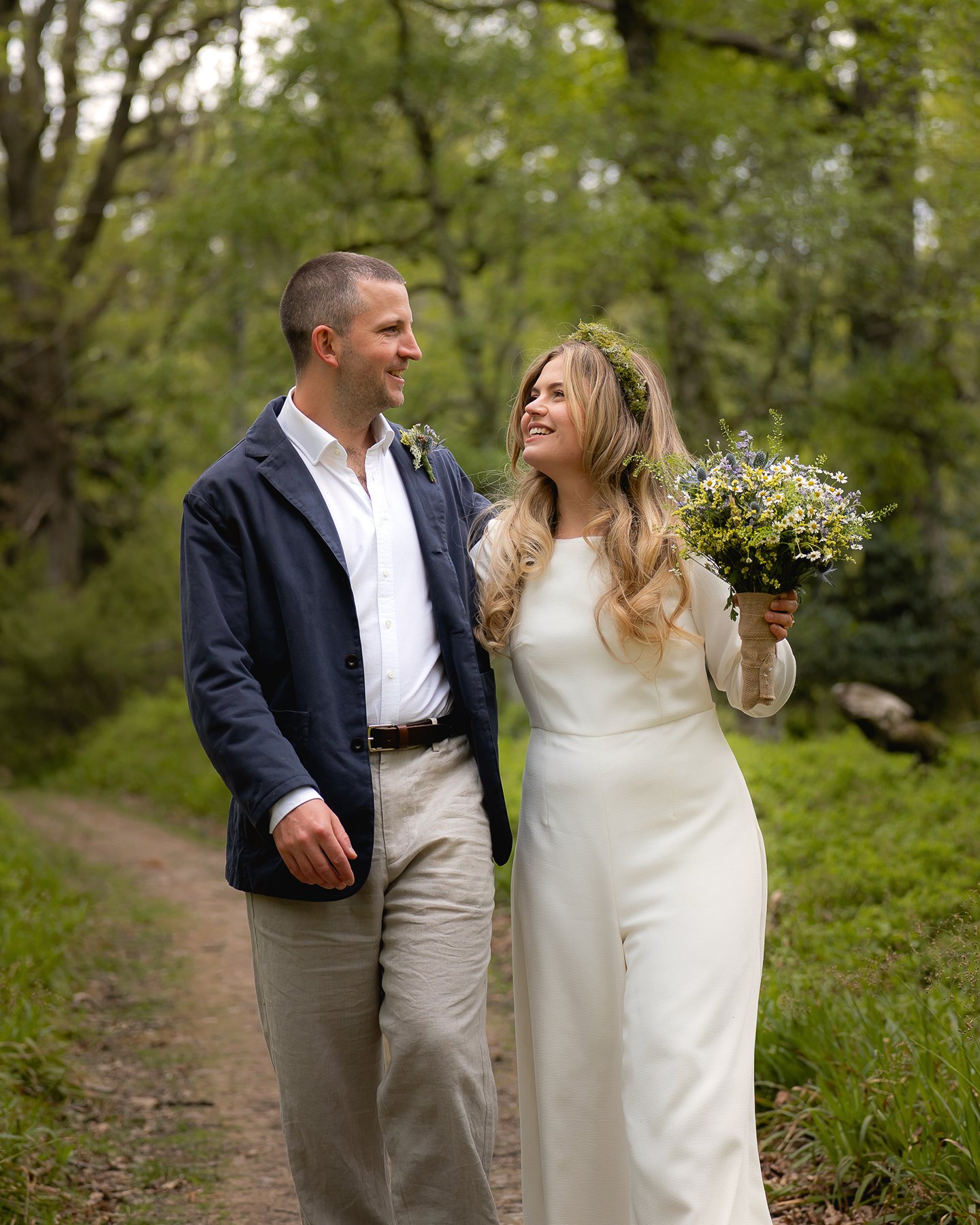 Smiling bride and groom walking together in woods, bride holding wildflower bouquet.