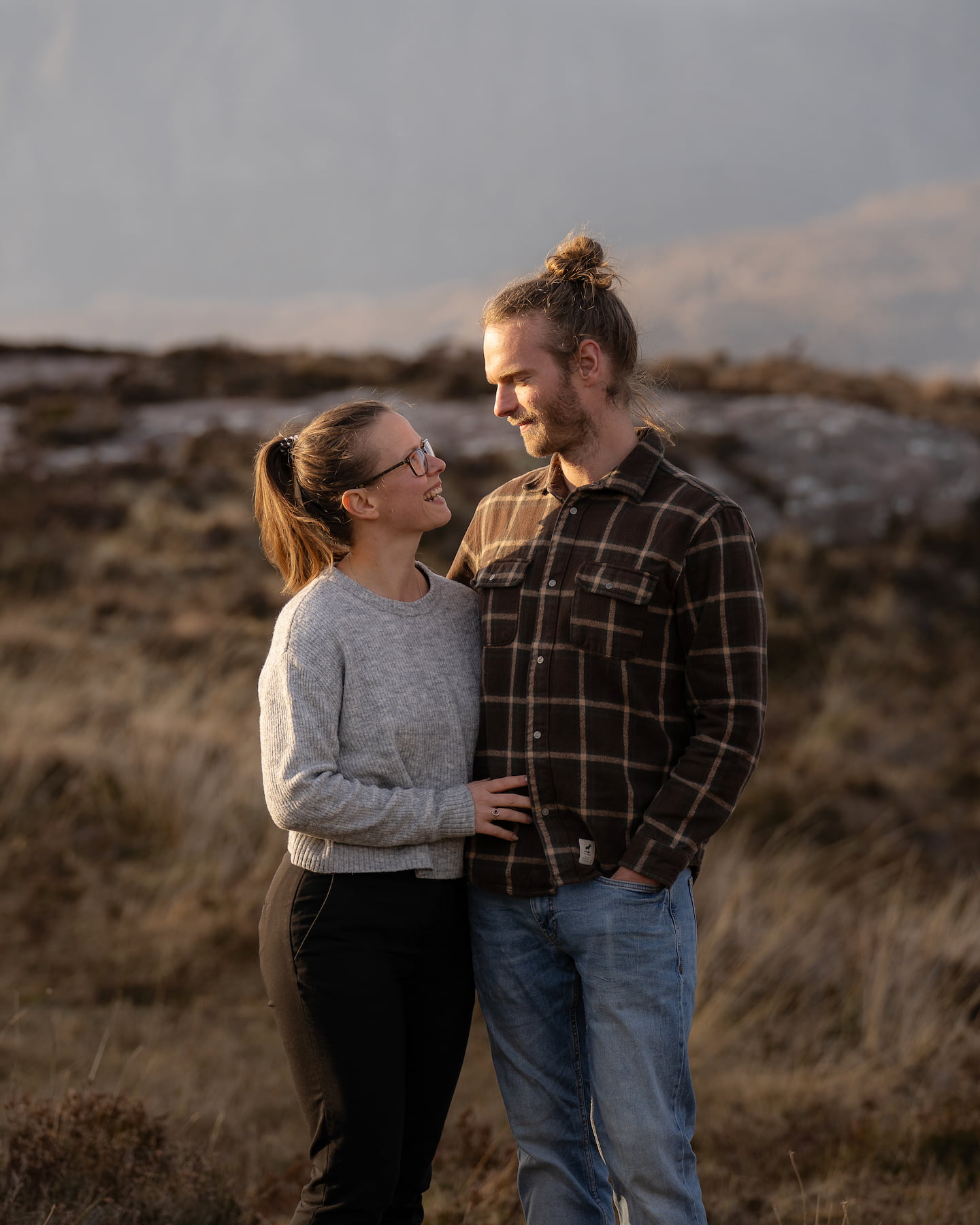 Couple smiling at each other, standing close in a mountain landscape, sharing an intimate moment outdoors.